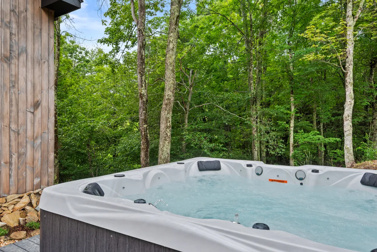 This exterior shot captures a private hot tub nestled beside a wooden cabin, surrounded by lush greenery. The scene exudes tranquility and exclusivity, with the hot tub positioned for privacy and relaxation, perfect for unwinding under the stars.
