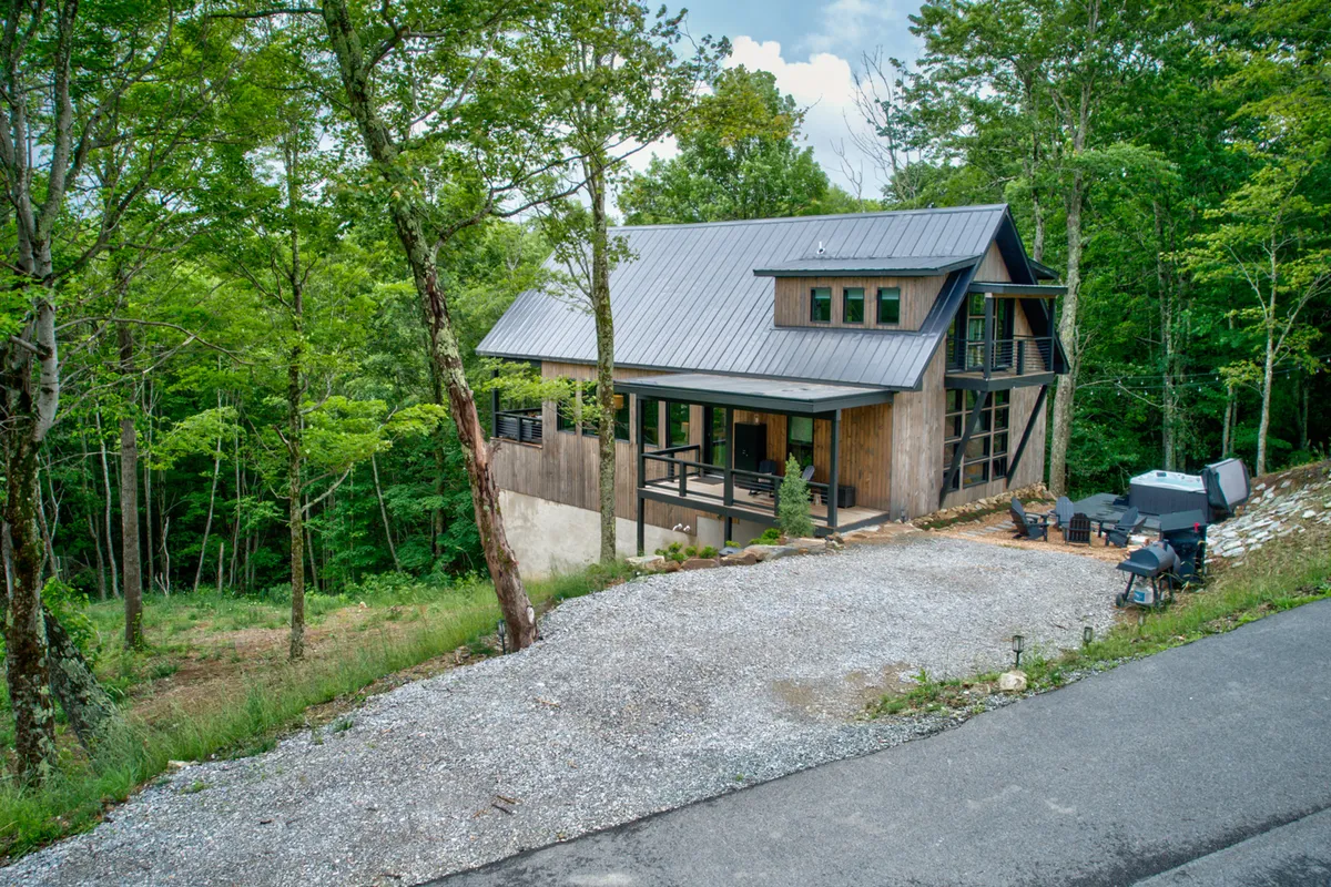 This image shows a modern cabin nestled among tall trees, with a gravel driveway leading up to the house and an inviting outdoor seating area on a spacious deck. The lush greenery and forested surroundings evoke a sense of privacy and immersion in nature, perfect for groups seeking a peaceful retreat. The setup hints at opportunities for outdoor gatherings and stargazing in the evenings.
