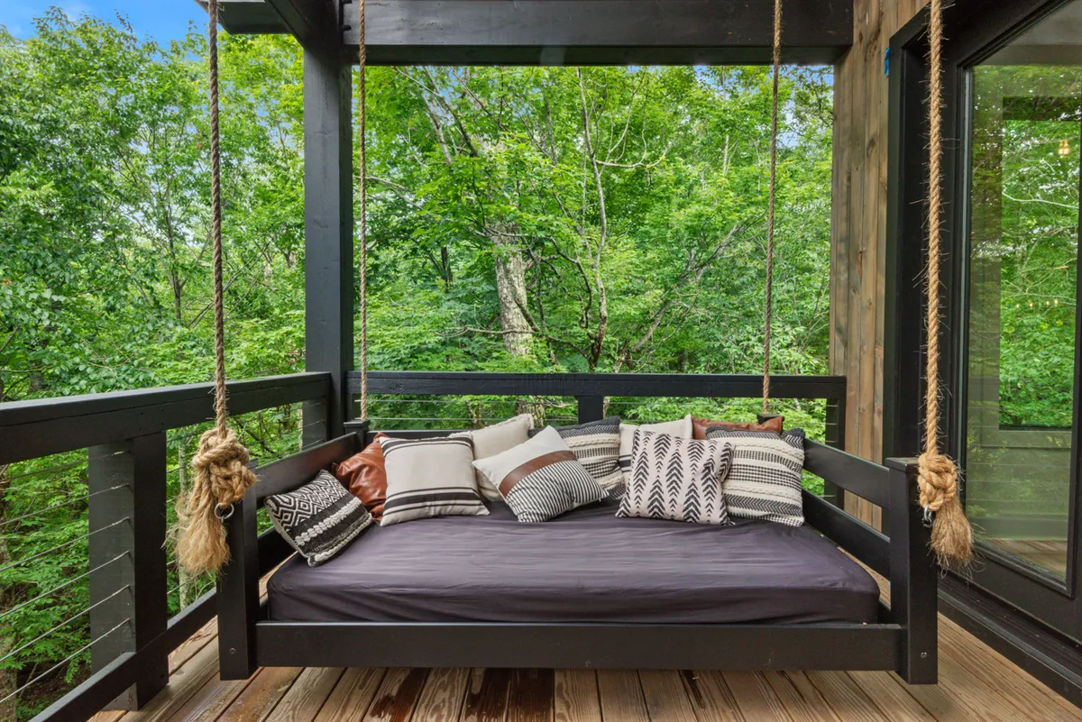 A suspended swinging daybed, adorned with decorative pillows, faces out toward lush greenery. The tranquil vibe and inviting setup suggest a perfect spot for quiet reflection, morning coffee, or stargazing.