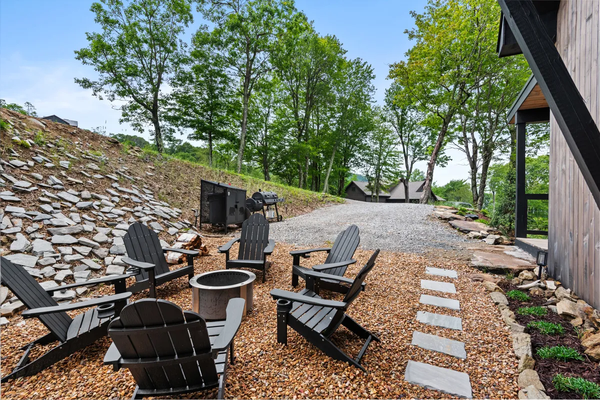 A cozy outdoor area with Adirondack chairs arranged around a modern fire pit and a grill area, all set along a gravel path and framed by dense trees. The scene is designed for social gatherings, stargazing, and enjoying the mountain air in comfort.