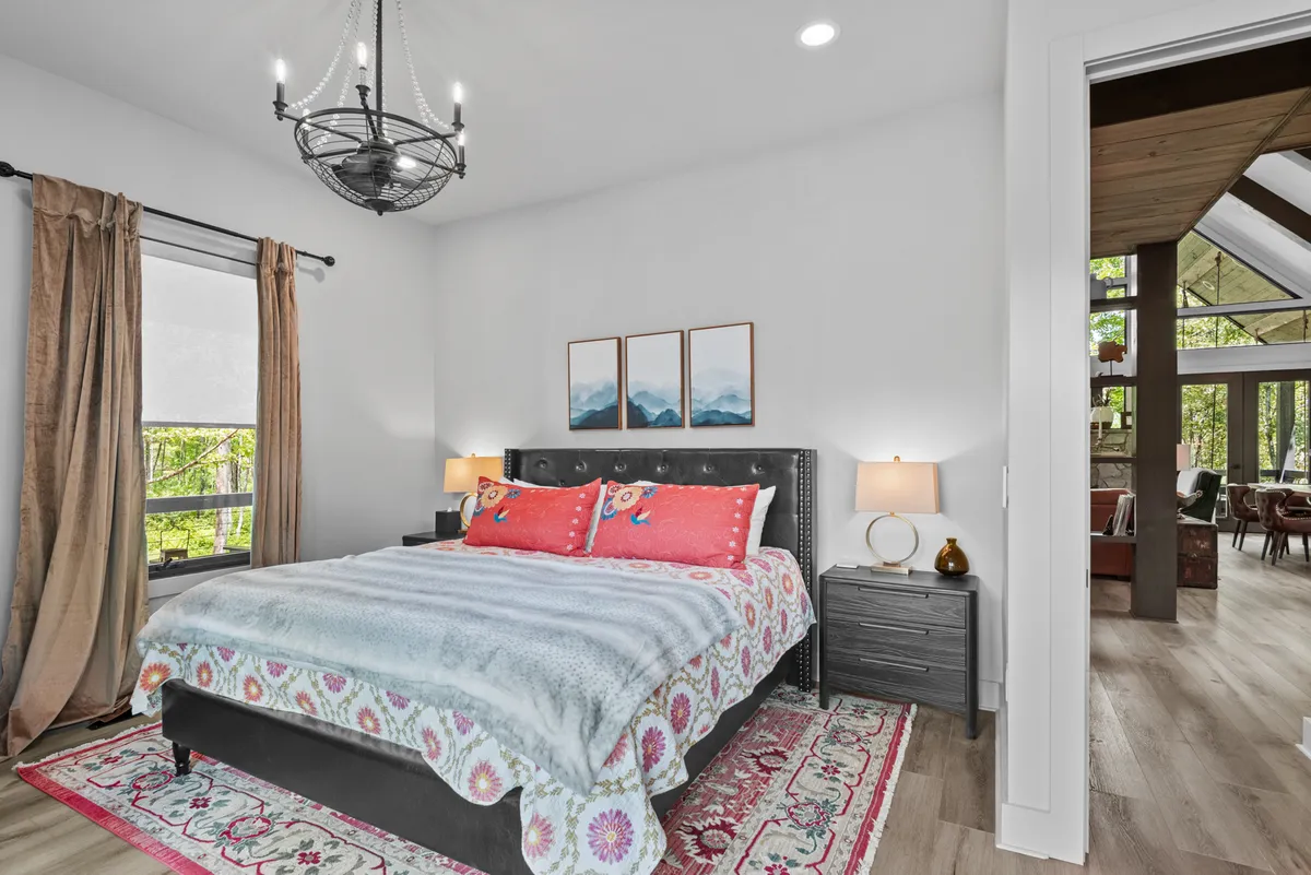 This interior image showcases a king-sized bed with colorful, crisp bedding, a chandelier overhead, and a large window that bathes the room in natural light. The ambiance is serene and comfortable, providing visual cues of a restful and luxurious retreat.