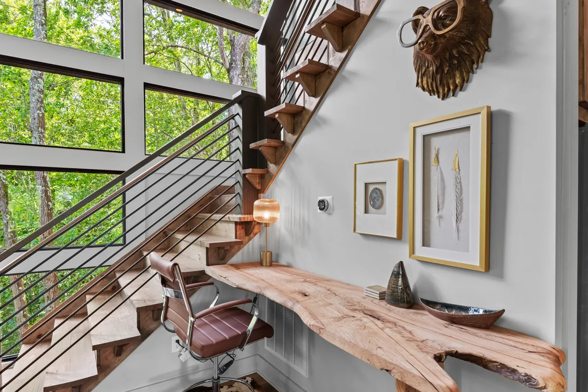 A creative nook on the main level featuring a wooden desk positioned by a large window, decorative wall art, and a unique animal mask. The space offers a view of a staircase and is filled with natural light, creating a peaceful setting ideal for working, reading, or enjoying a coffee while gazing at the surrounding nature.