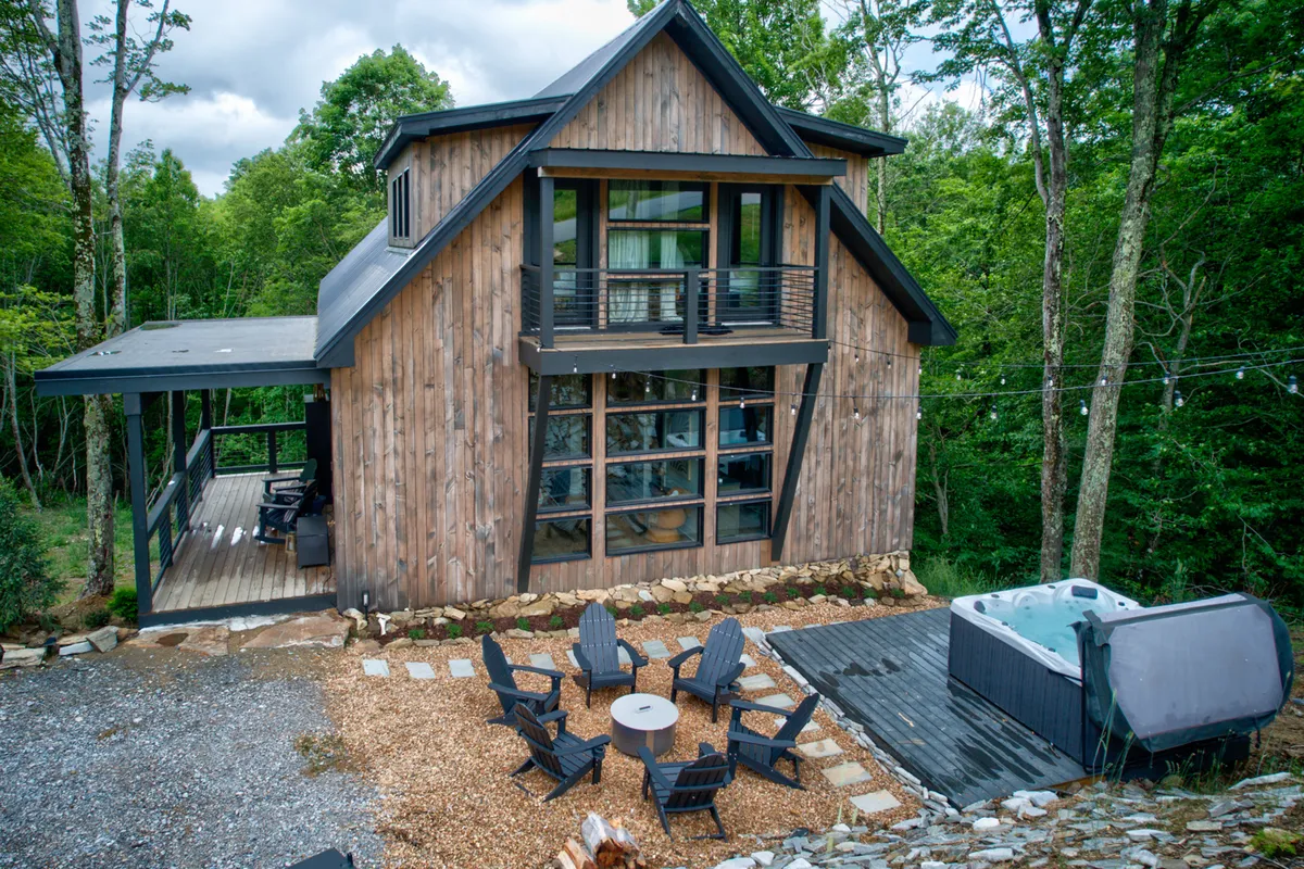 A close-up exterior view of the modern cabin's sleek structure with an inviting outdoor seating area, a bubbling hot tub, and tall trees encircling the space. The setting feels cozy yet upscale, hinting at new construction and thoughtful amenities.