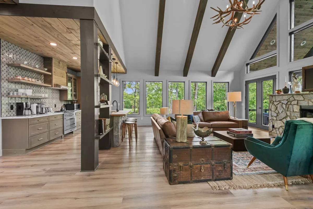 A wide-angle view of a spacious open-concept interior featuring a plush sofa facing a modern stone fireplace, a dining area with a wood table and stylish chairs, and a contemporary kitchen with granite countertops and stainless steel appliances. Large windows bathe the space in sunlight and offer views of the natural surroundings. The room is accented with exposed wood beams and a mix of modern and mid-century furnishings, creating a welcoming yet chic mountain retreat.
