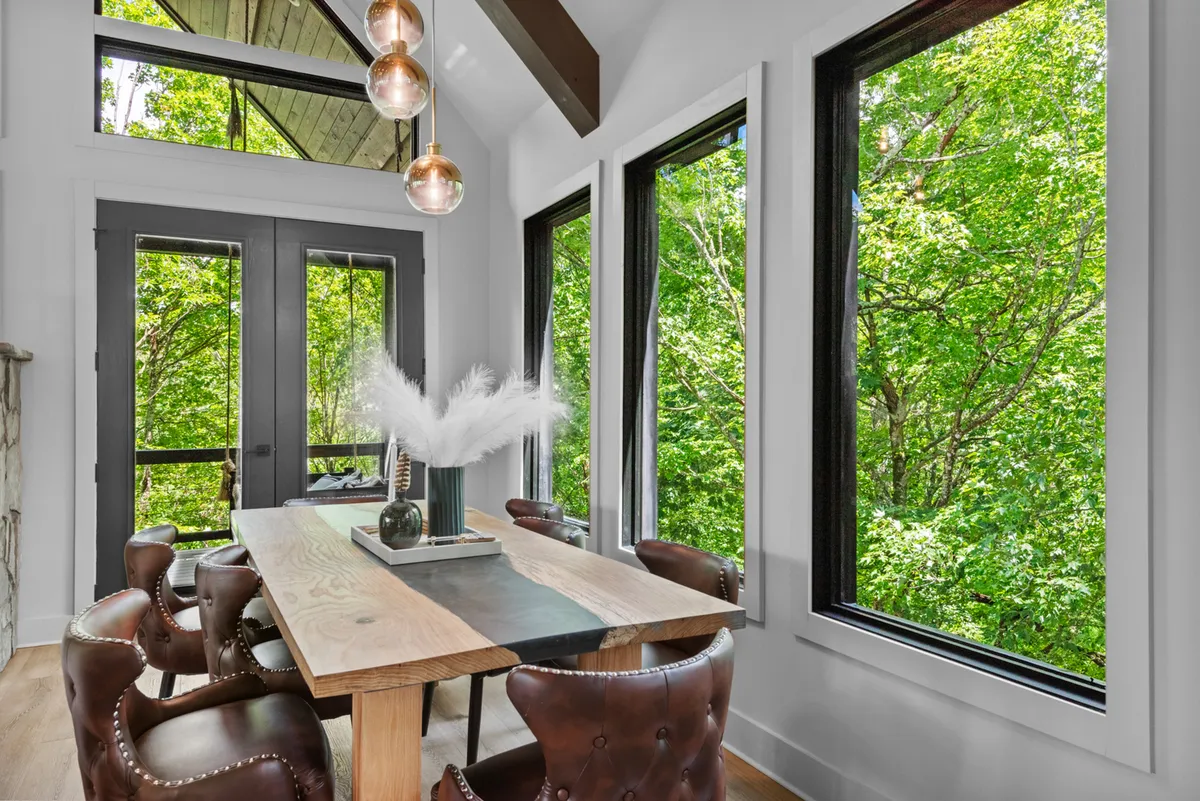 An interior shot focusing on a stunning wooden dining table beneath a chandelier, flanked by large windows that open onto lush greenery. The space feels both welcoming and sophisticated, with abundant natural light and peaceful views.