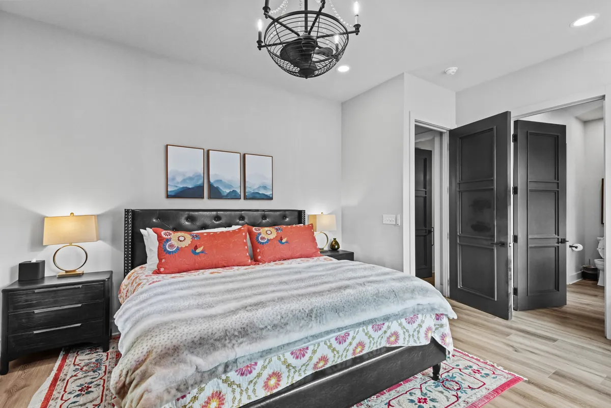 An inviting king bedroom with colorful bedding, nightstands with lamps, framed artwork, and an open door leading to another room. The scene is well-lit with natural light, accentuating the calming and welcoming mood.