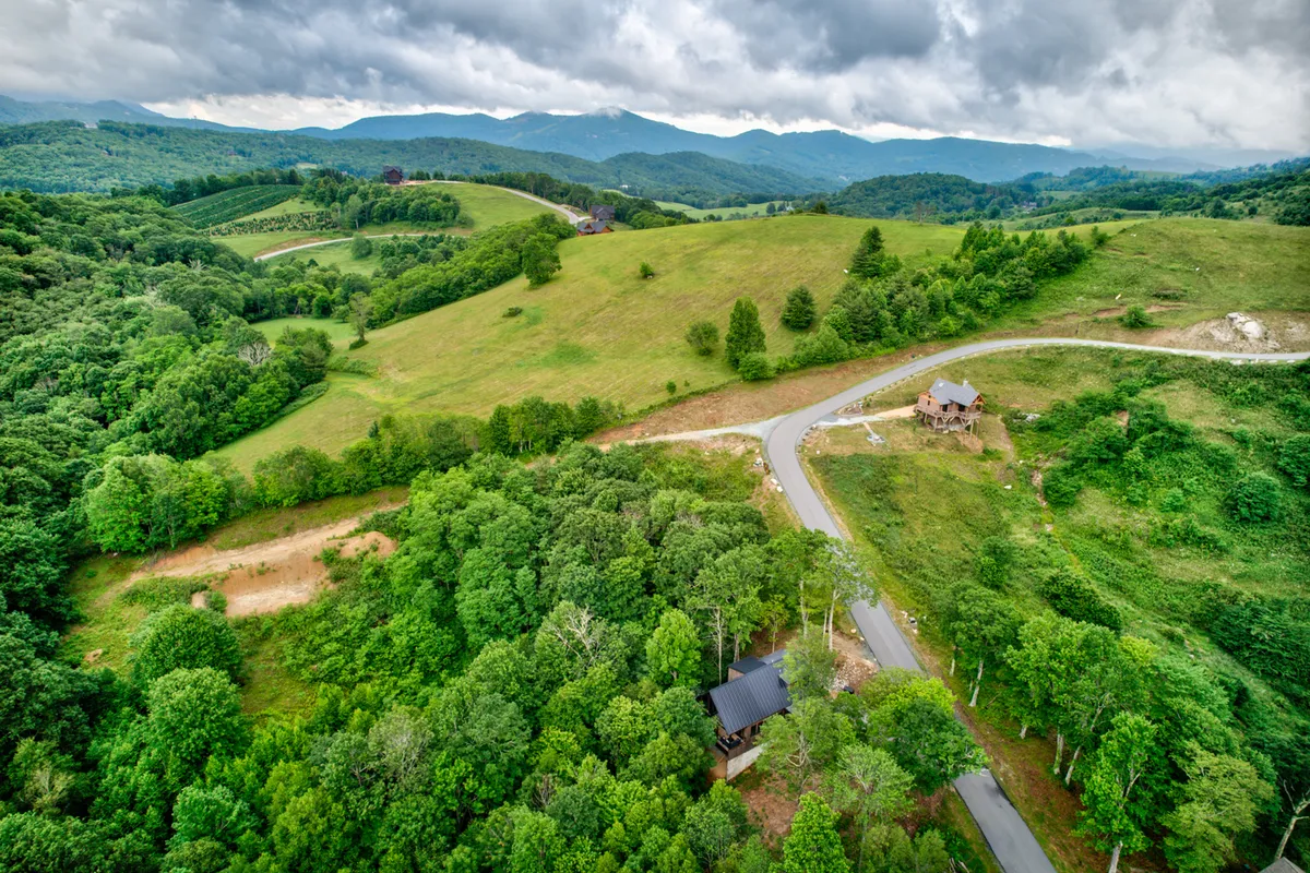 An expansive view of rolling hills and dense greenery, with a winding road leading toward distant mountains. Scattered houses are visible amidst the lush landscape, suggesting easy access to local trails and scenic outdoor adventures.