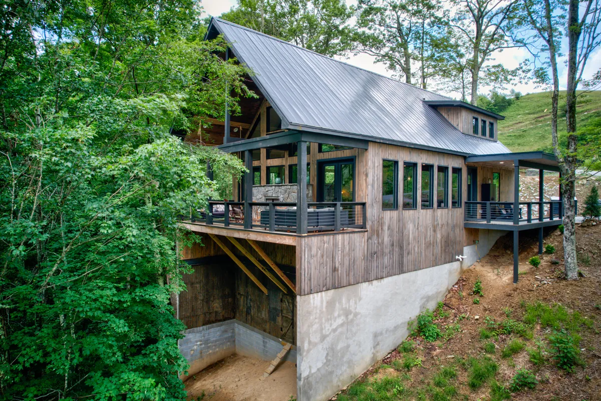 A wide view of the home’s wooden exterior, featuring large windows and a spacious deck with outdoor seating, all nestled among tall trees. The setting invites guests to enjoy morning coffee, sunset views, and the natural beauty of the surrounding mountains.