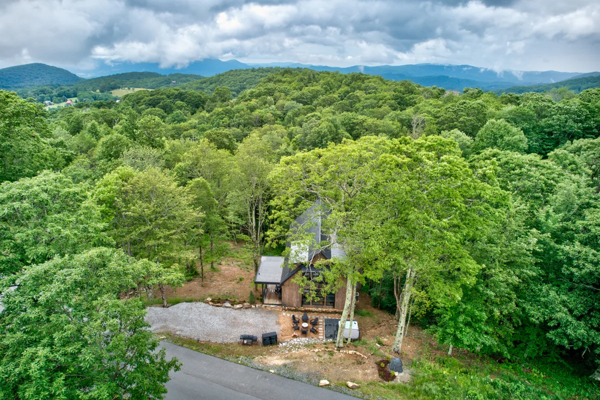 A wide shot of the modern mountain home nestled among mature trees, with the broad mountain range visible in the background. The scene is bathed in peaceful natural light, evoking a sense of seclusion and tranquility.