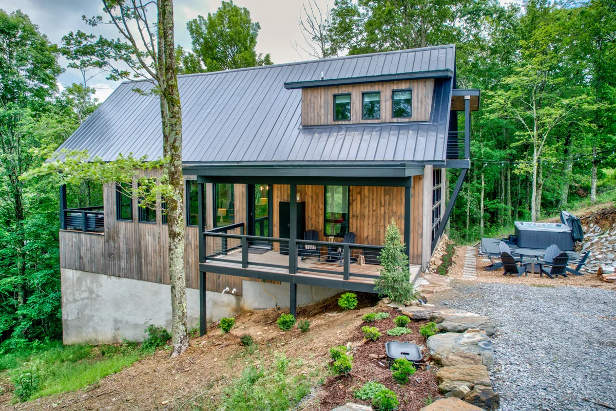 The image showcases the exterior of a modern mountain home nestled in Banner Elk, North Carolina. The house features striking contemporary architecture with large windows, a spacious deck outfitted for gatherings, and a bubbling hot tub overlooking a landscaped garden. The setting radiates tranquility, inviting guests to relax outdoors and take in the surrounding natural beauty.