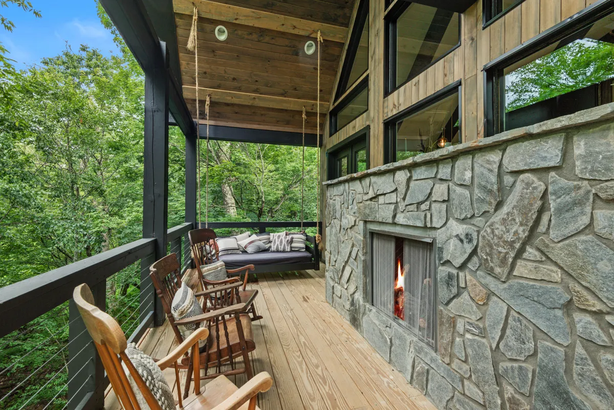 A tranquil deck scene centered around a rustic rock fireplace, with a swing seat and wooden rocking chairs arranged for relaxation. Large windows in the background connect the space to the indoors, while the natural setting emphasizes comfort and cozy evenings by the fire.