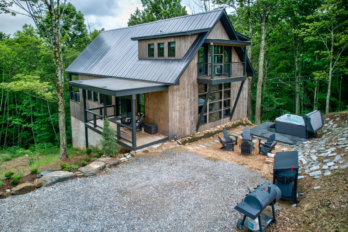 This image displays a modern cabin structure with a spacious deck, featuring both a hot tub and a grill area. The deck is surrounded by lush greenery, and the scene captures an inviting outdoor atmosphere ideal for gatherings and relaxation. The grill and hot tub are clearly visible, offering a glimpse of the luxurious amenities mentioned.
