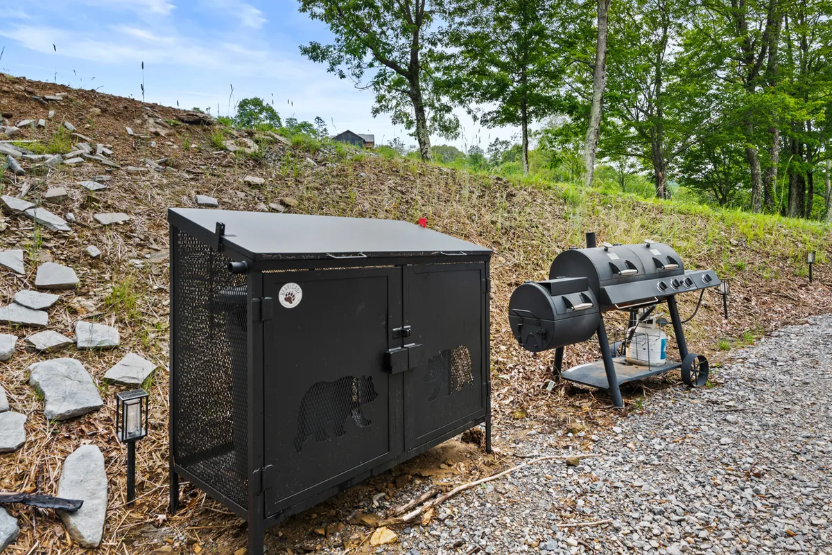 The image focuses on an exterior barbecue grill area featuring a bear-proof trash container, set against a backdrop of natural greenery. The scene suggests outdoor cooking and relaxed meals, as well as the consideration given to local wildlife. The area appears inviting for group cookouts and casual dining after a day of adventure.